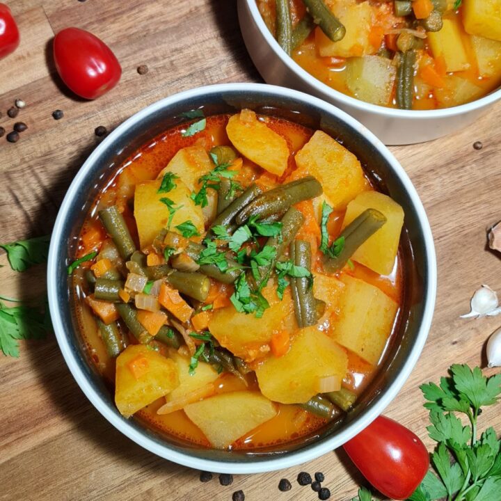 A bowl of hearty potato and green bean soup garnished with fresh parsley, surrounded by ingredients like cherry tomatoes, garlic, and peppercorns on a wooden surface.