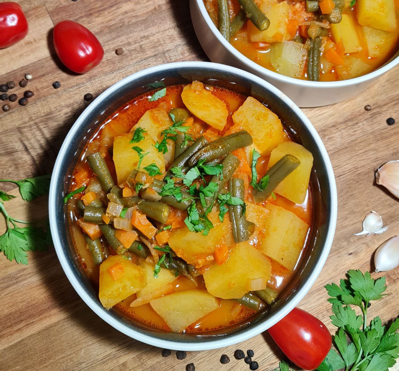 A bowl of hearty potato and green bean soup garnished with fresh parsley, surrounded by ingredients like cherry tomatoes, garlic, and peppercorns on a wooden surface.