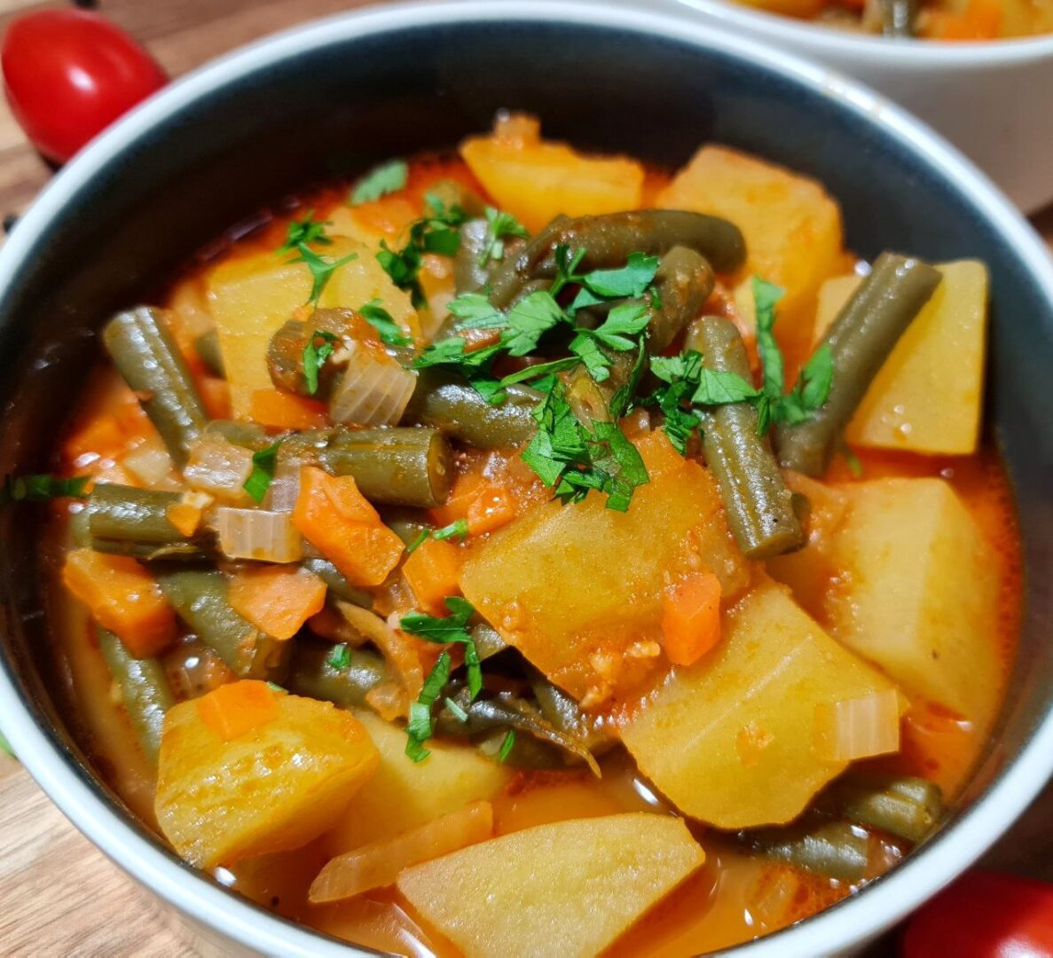  A wooden board with two bowls of potato and green beans soup, garnished with fresh parsley, surrounded by cherry tomatoes, garlic cloves, black peppercorns, and parsley sprigs.