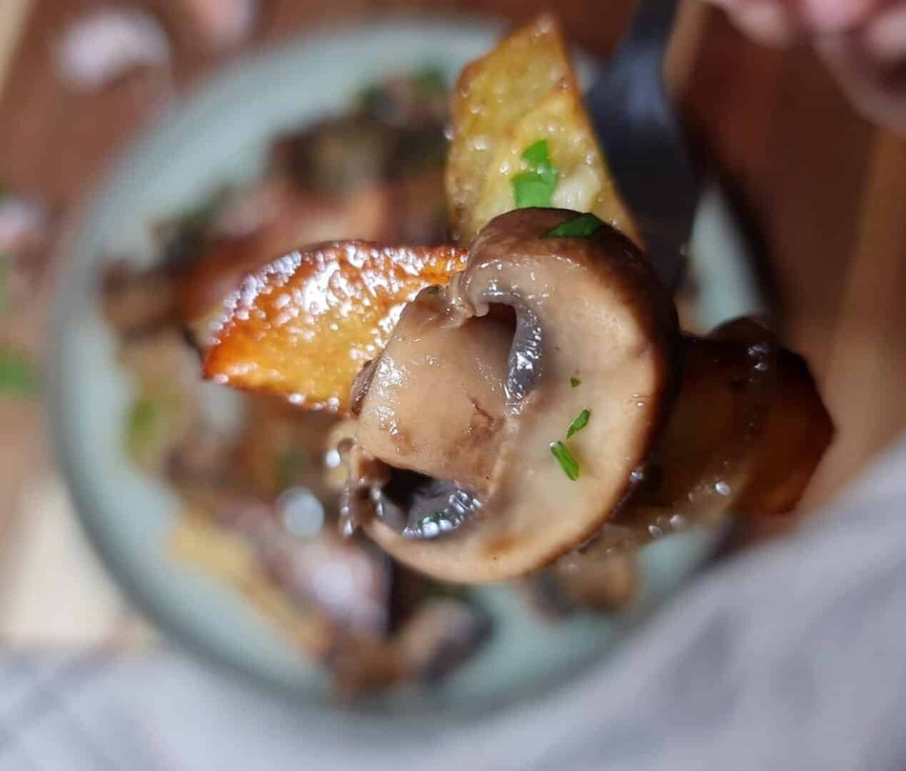 Close-up of a fork holding a piece of roasted potato and mushroom garnished with green herbs.