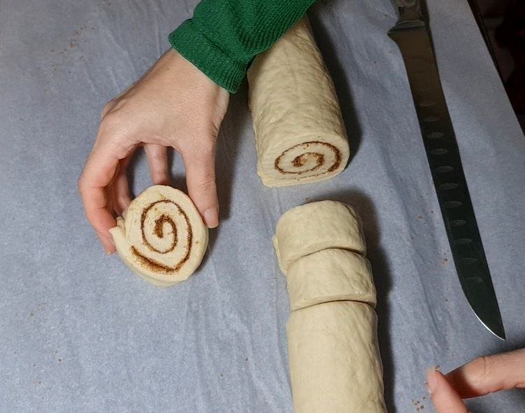 Hands preparing vegan cinnamon rolls by slicing dough into individual rolls on a parchment-lined surface.