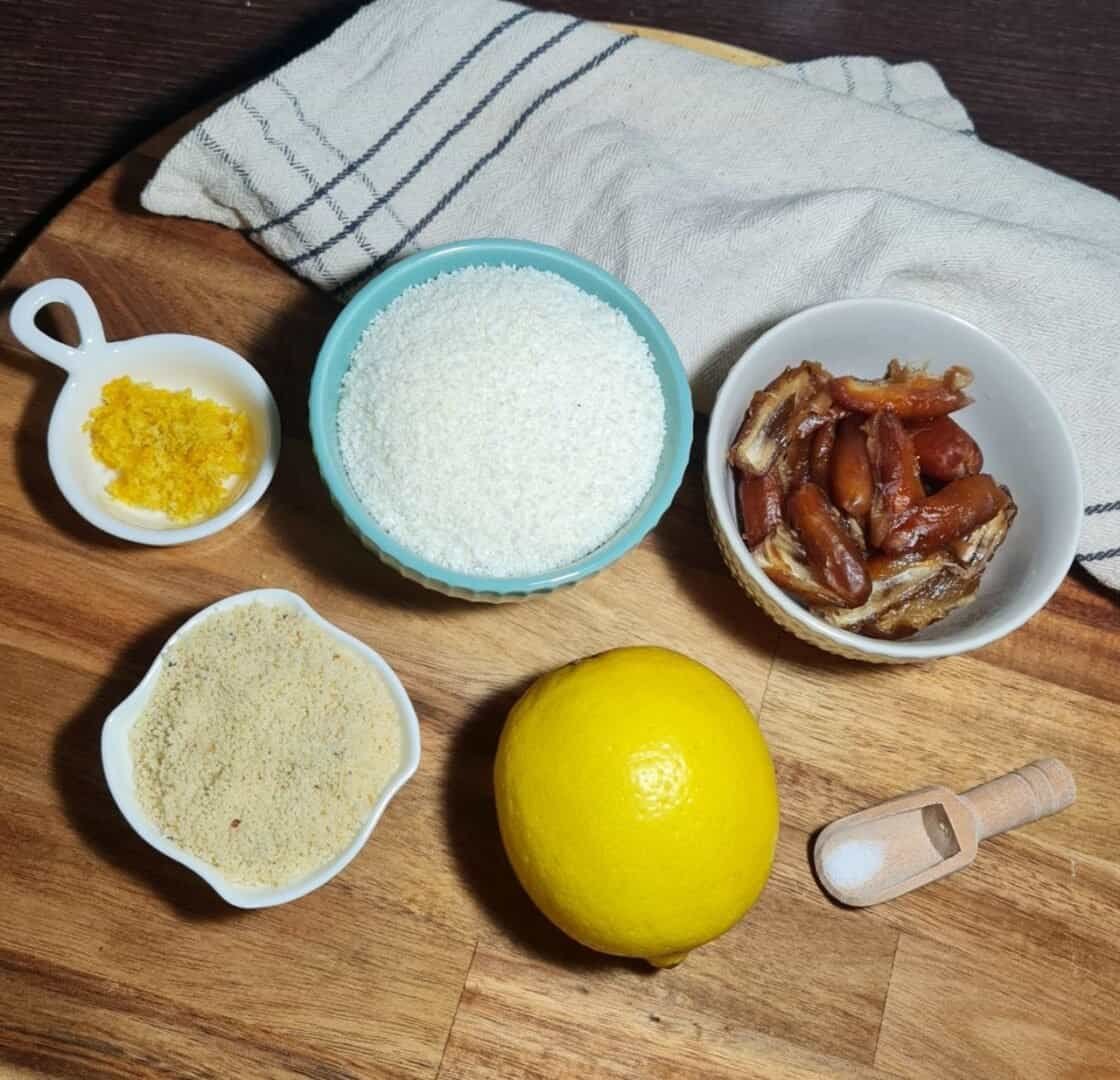 Ingredients for vegan lemon coconut balls including lemon zest, shredded coconut, dates, almond flour, a lemon, and a pinch of salt on a wooden board.