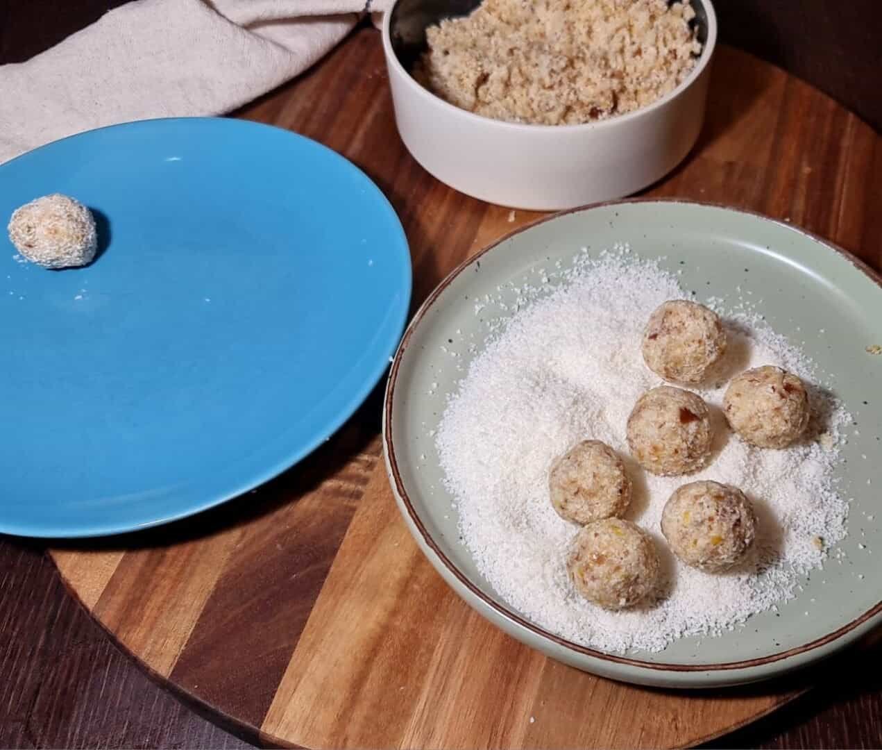 A preparation scene of vegan lemon coconut balls, showing the rolling process with a blue plate, a green plate with shredded coconut, and a bowl of the mixture.