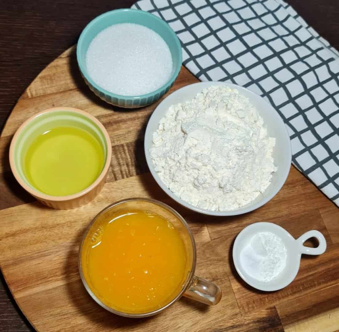  Ingredients for vegan orange cake including flour, sugar, orange juice, oil, and baking powder arranged on a wooden board.