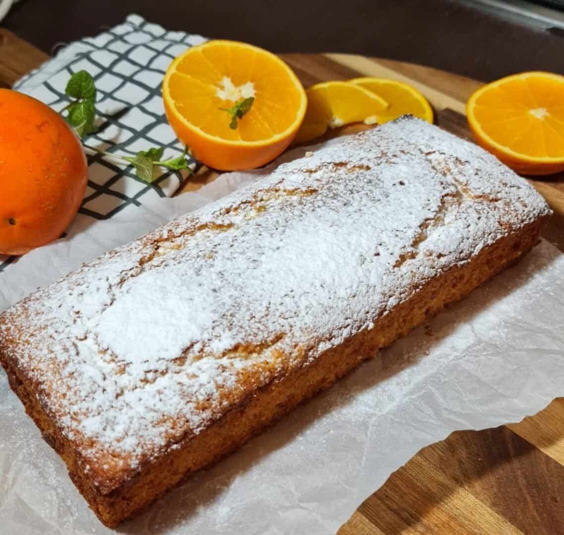 Vegan orange cake dusted with powdered sugar, surrounded by fresh oranges and mint leaves on a wooden board.