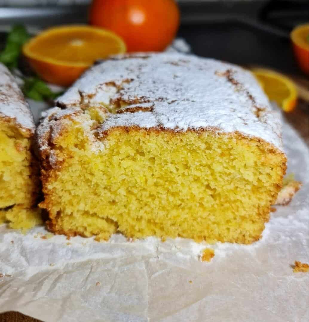 A close-up of a freshly baked vegan orange cake topped with powdered sugar, with fresh oranges in the background.