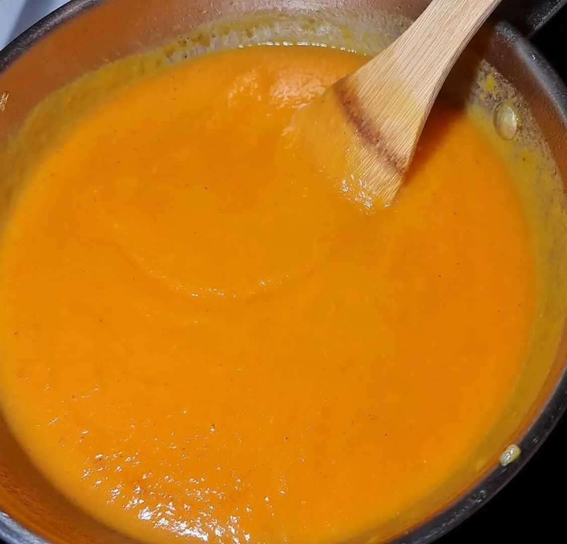 A pot of smooth, vibrant orange carrot soup being stirred with a wooden spoon.