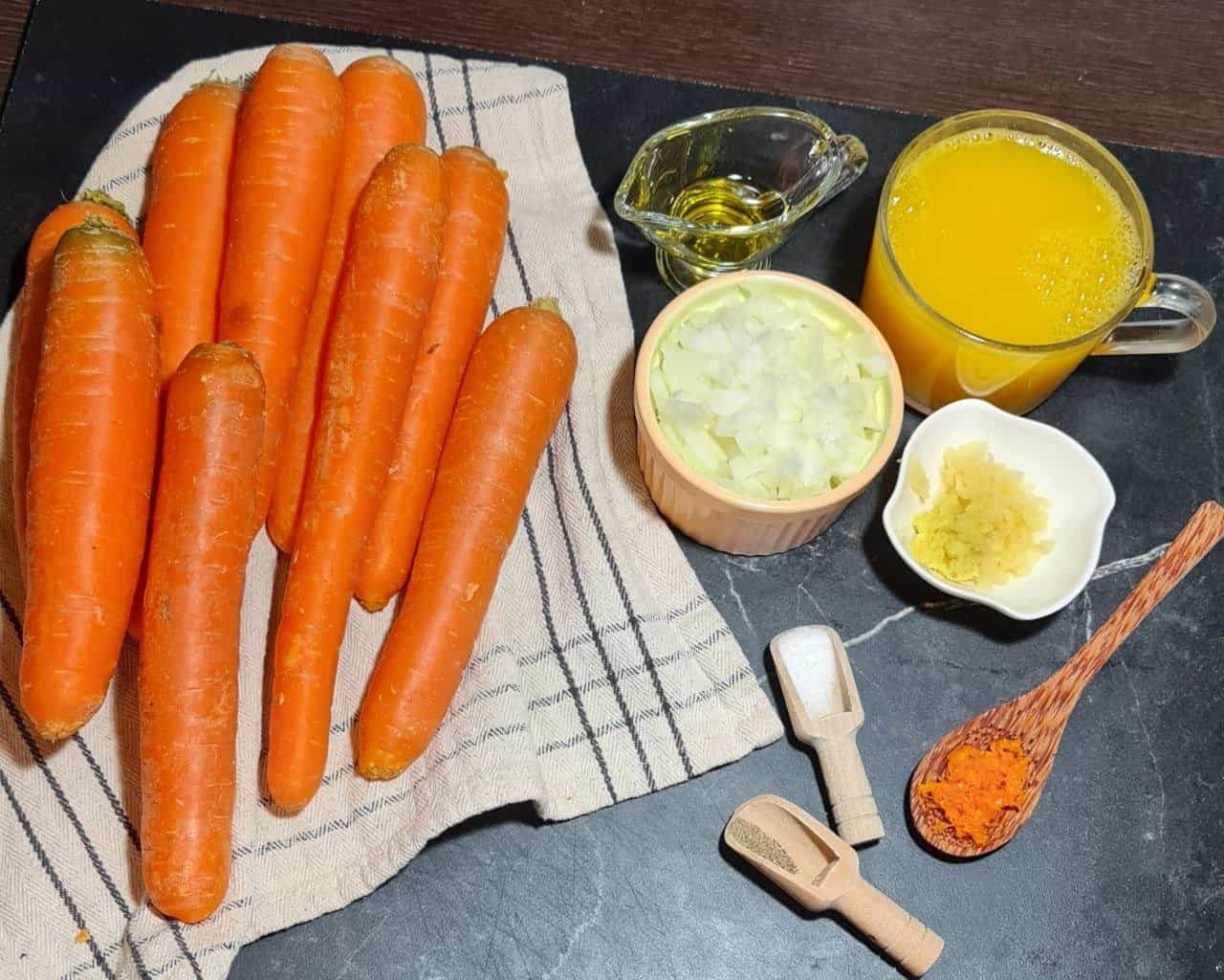 Ingredients for vegan orange carrot soup including fresh carrots, chopped onions, minced ginger, turmeric, salt, pepper, olive oil, and orange juice.