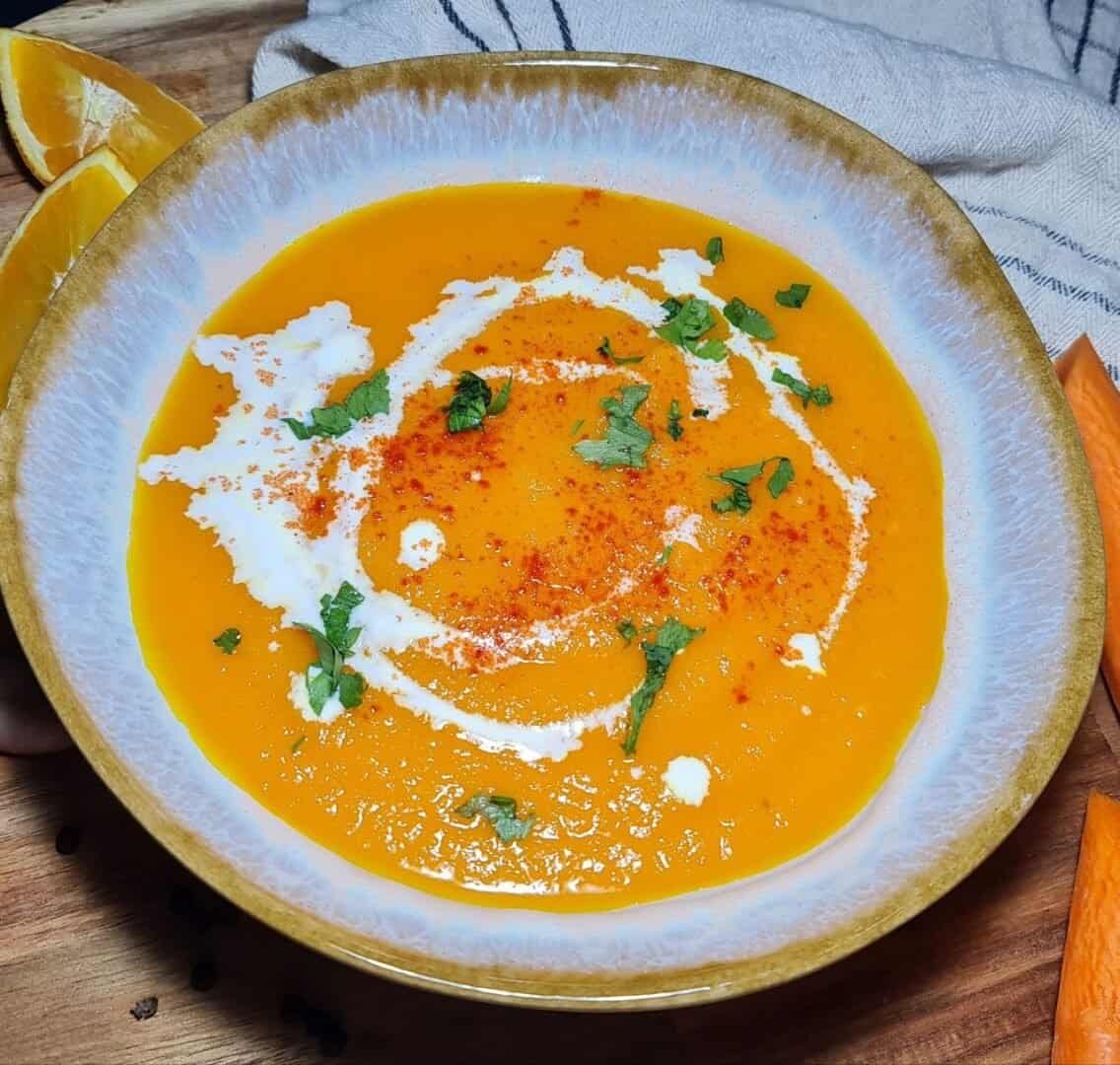 A bowl of vibrant orange carrot soup garnished with cream, cilantro, and spices, placed on a wooden board with fresh carrot pieces and orange slices.