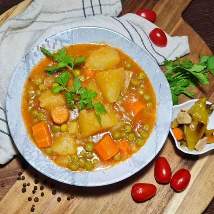 A bowl of vegan peas and potato stew garnished with fresh parsley, featuring chunks of potatoes, carrots, and peas in a rich tomato-based broth.