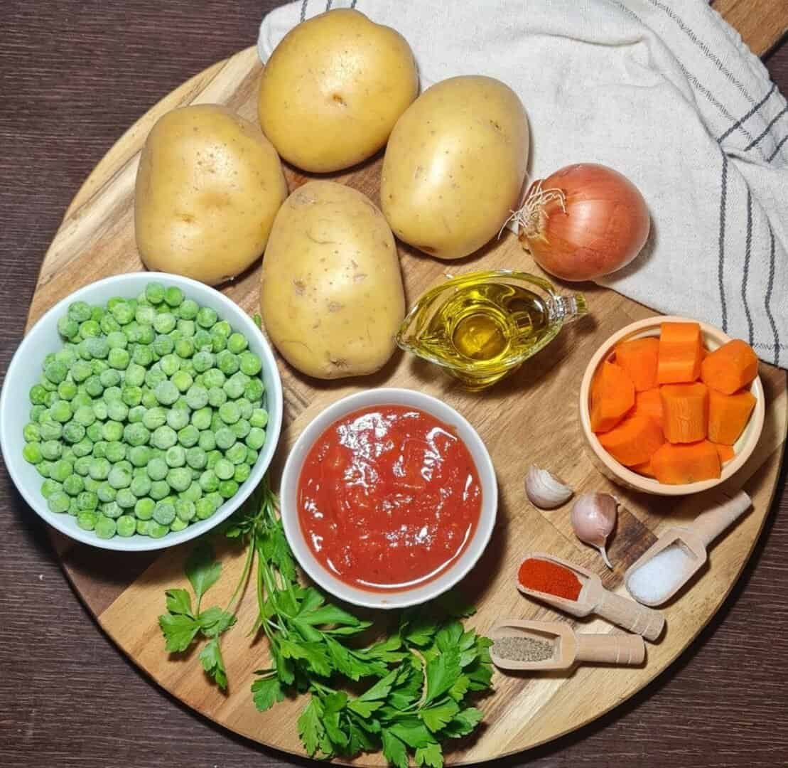  Ingredients for vegan peas and potato stew including potatoes, peas, carrots, onion, garlic, tomato sauce, olive oil, parsley, and spices on a wooden board.