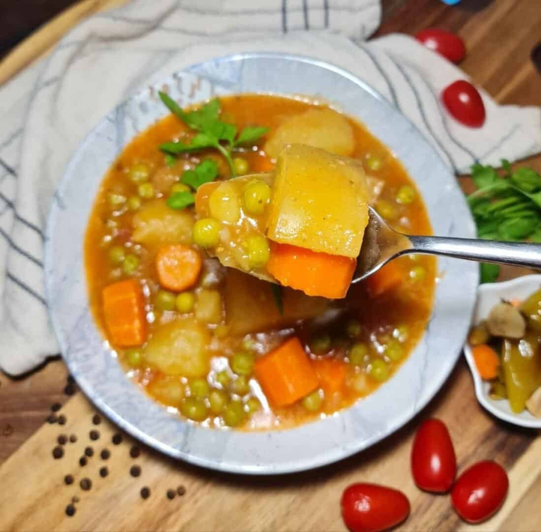 A close-up of a bowl of vegan peas and potato stew with a spoon lifting a portion of the stew, showcasing chunks of potatoes, carrots, and green peas in a rich broth.