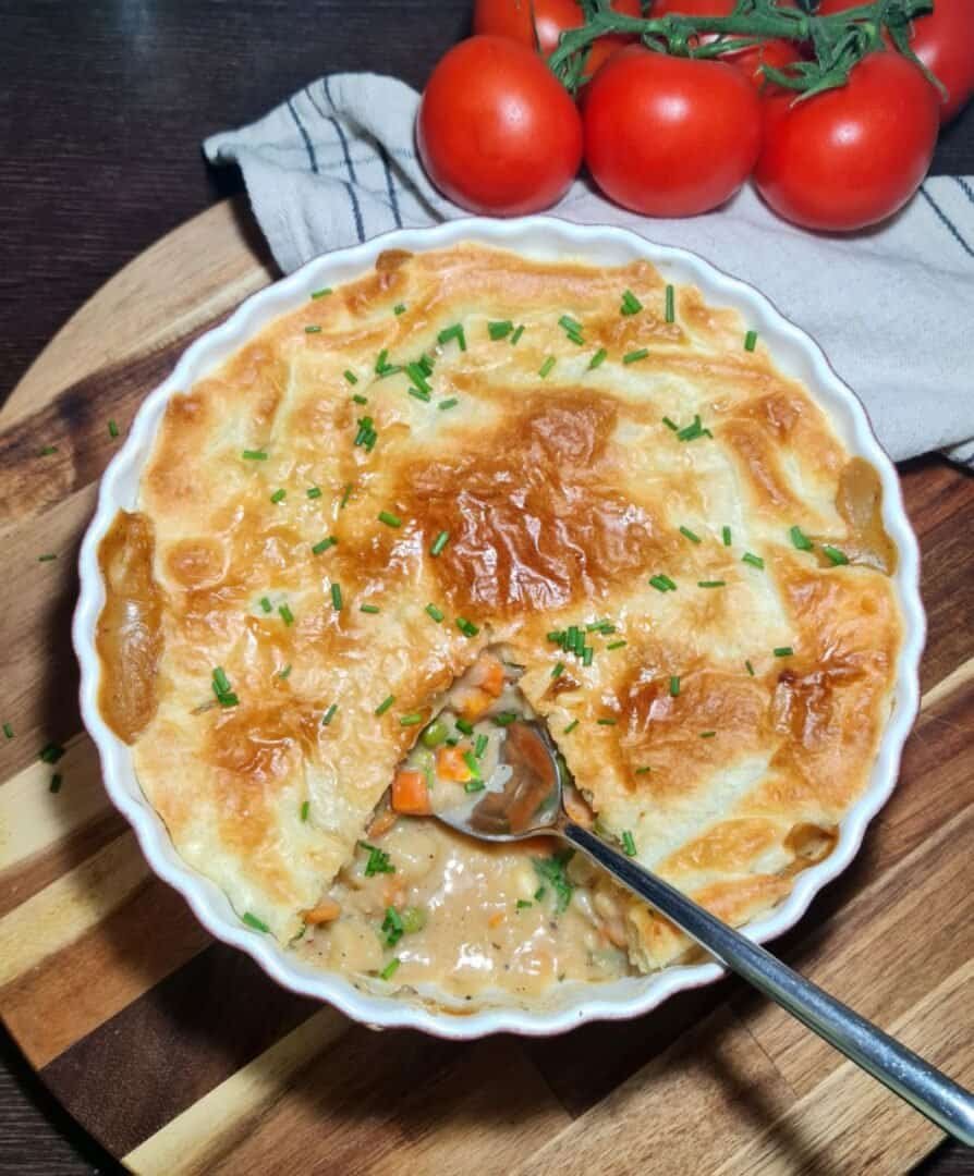 A freshly baked vegan pot pie with a golden-brown crust, garnished with chopped chives, and a spoon revealing the creamy vegetable filling inside.