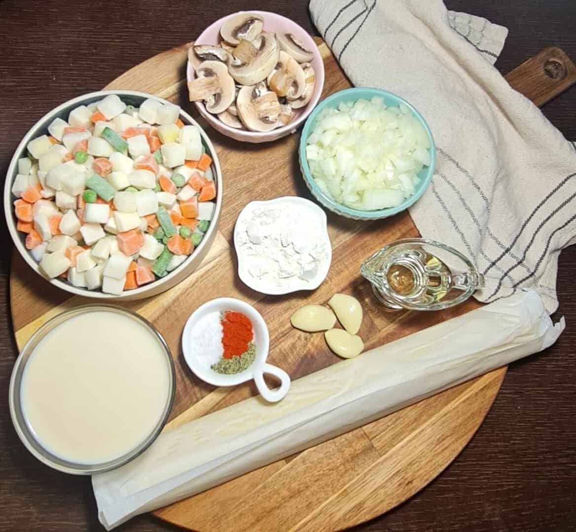 Ingredients for vegan pot pie including mixed vegetables, mushrooms, onions, garlic, flour, spices, oil, and plant-based milk arranged on a wooden board.