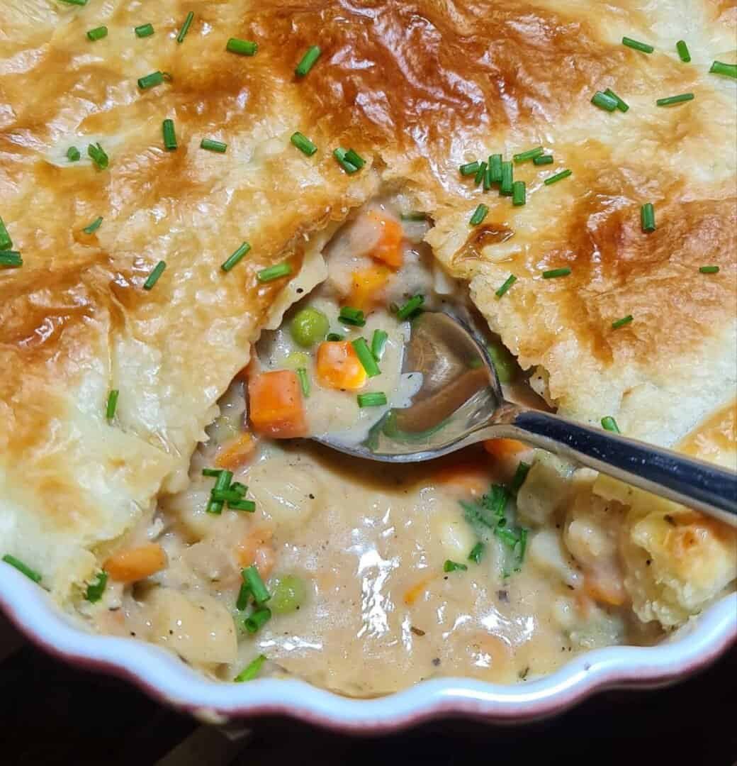 close-up of a vegan pot pie with a golden-brown crust, filled with creamy vegetables like carrots, peas, and potatoes, garnished with chopped chives.