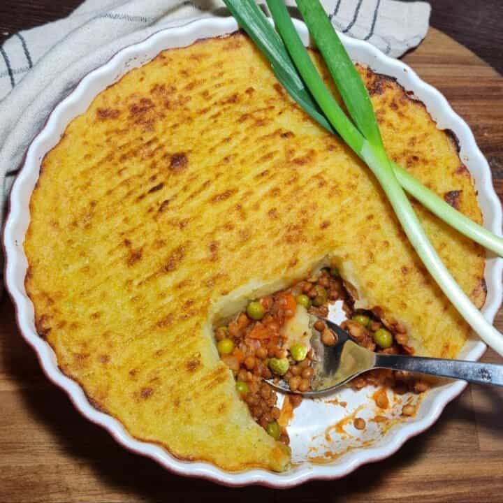 A freshly baked vegan shepherd's pie with a golden-brown mashed potato topping, garnished with green onions, and a serving spoon revealing the colorful vegetable filling inside.