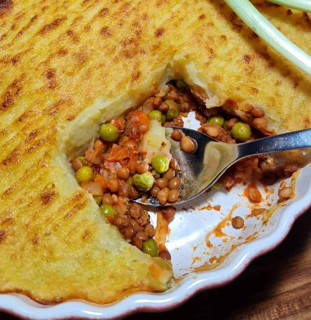 A close-up of a vegan shepherd's pie with a golden-brown mashed potato crust, revealing a filling of lentils, peas, and carrots.