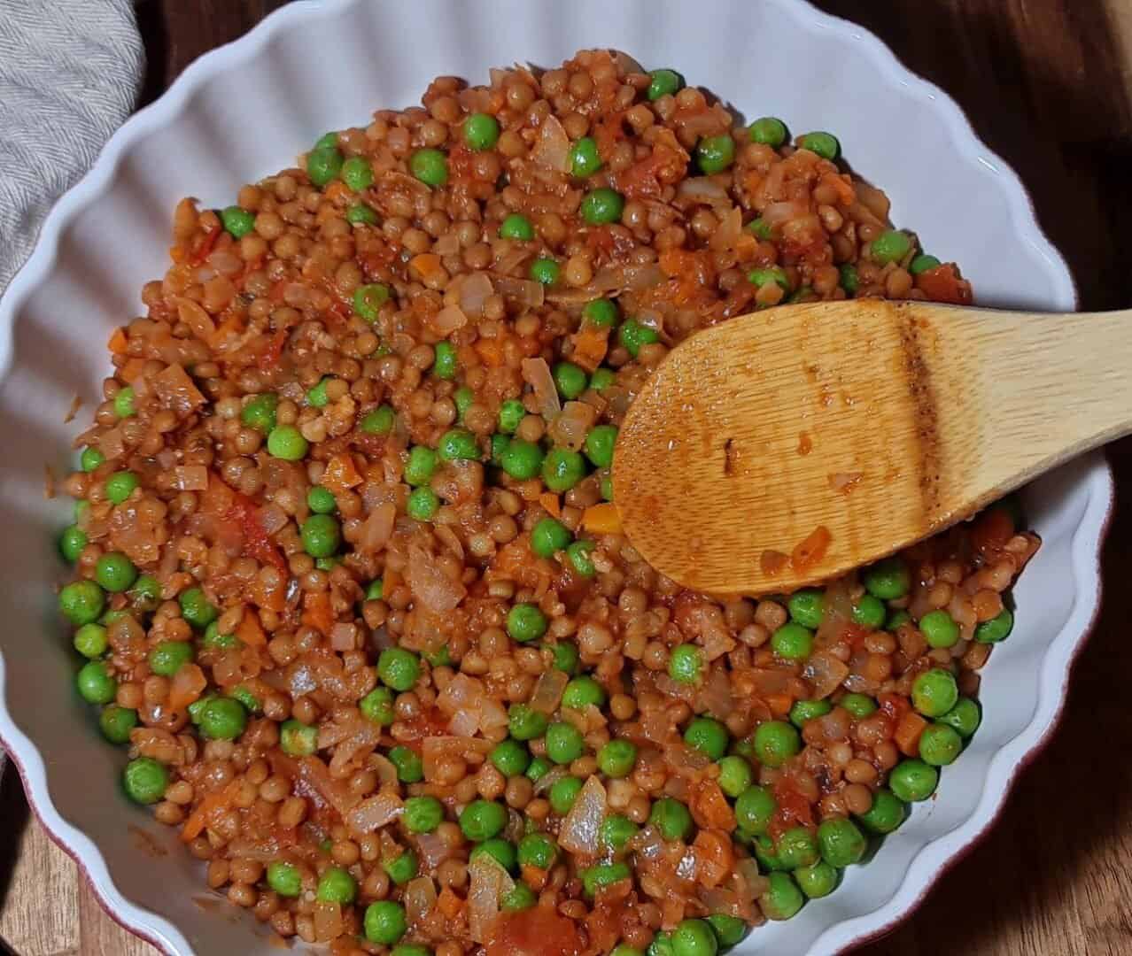 Close-up of vegan shepherd's pie filling with lentils, peas, and vegetables in a white dish with a wooden spoon.