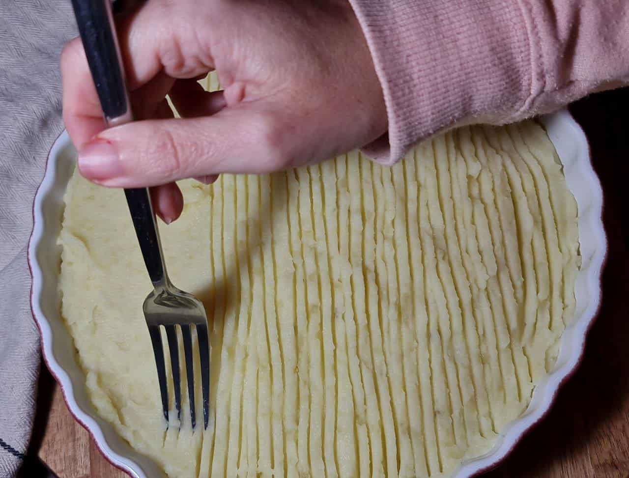 A hand using a fork to create lines on the mashed potato topping of a vegan shepherd's pie in a white baking dish.