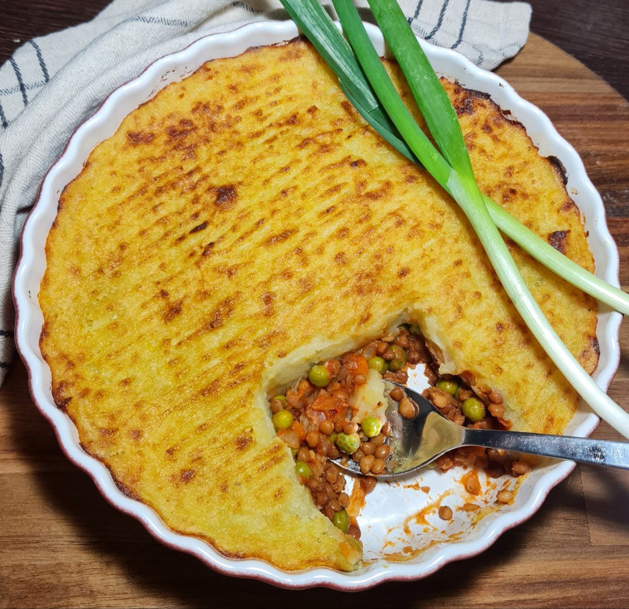 A freshly baked vegan shepherd's pie with a golden-brown mashed potato topping, garnished with green onions, and a serving spoon revealing the colorful vegetable filling inside.