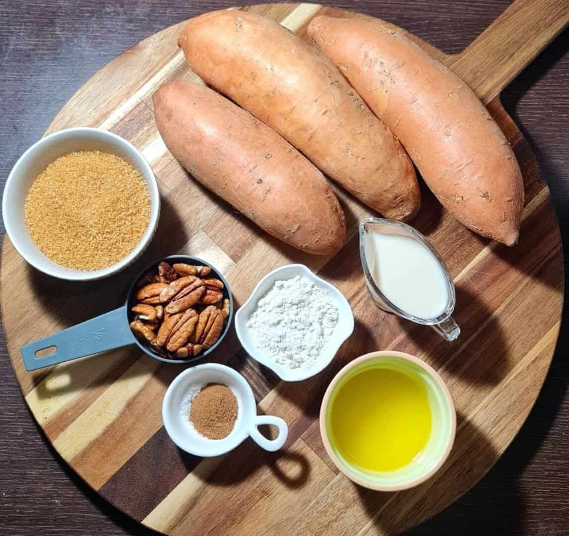  Ingredients for vegan sweet potato casserole including sweet potatoes, brown sugar, pecans, flour, plant-based milk, melted vegan butter, and spices arranged on a wooden board.