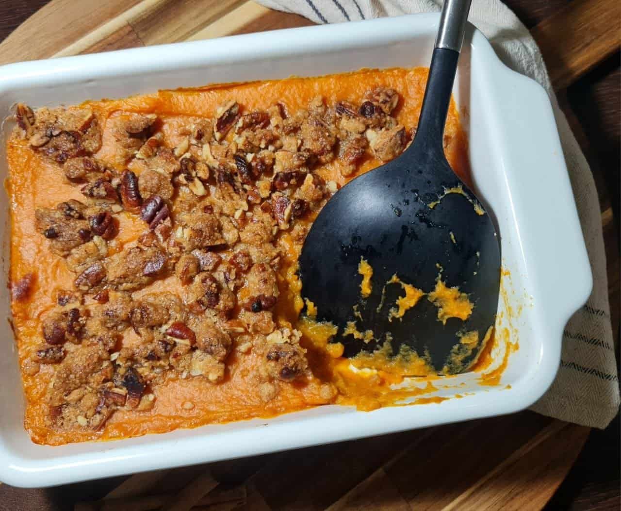 A close-up of a vegan sweet potato casserole in a white baking dish, topped with a crunchy pecan and oat crumble, with a black serving spoon resting inside.