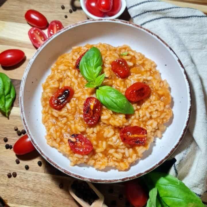 A bowl of vegan tomato risotto garnished with fresh basil leaves, surrounded by cherry tomatoes, black peppercorns, and basil leaves on a wooden board.