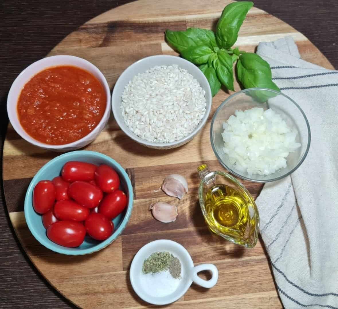 Ingredients for vegan tomato risotto including tomato sauce, rice, cherry tomatoes, chopped onions, garlic cloves, olive oil, basil leaves, and seasonings on a wooden board.