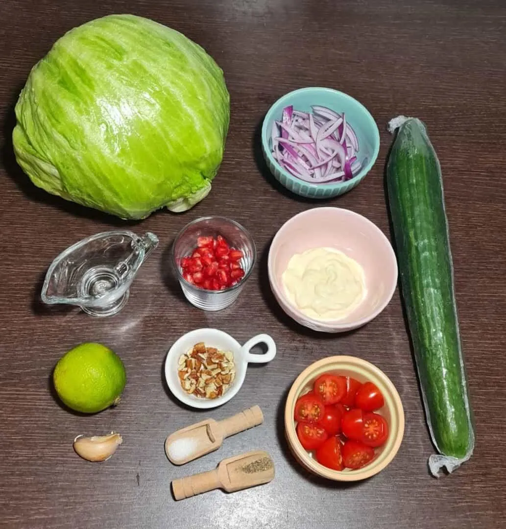 Ingredients for a vegan wedge salad including iceberg lettuce, cucumber, cherry tomatoes, red onion, pomegranate seeds, mayonnaise, lime, garlic, walnuts, salt, and pepper.