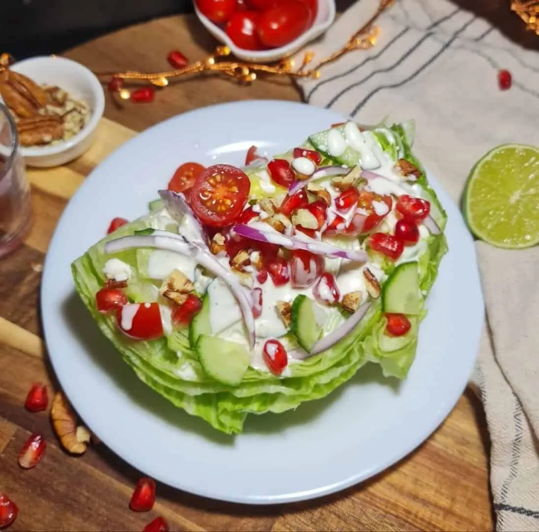 A vibrant vegan wedge salad topped with cherry tomatoes, cucumber slices, red onion, pomegranate seeds, and a creamy dressing, served on a white plate.