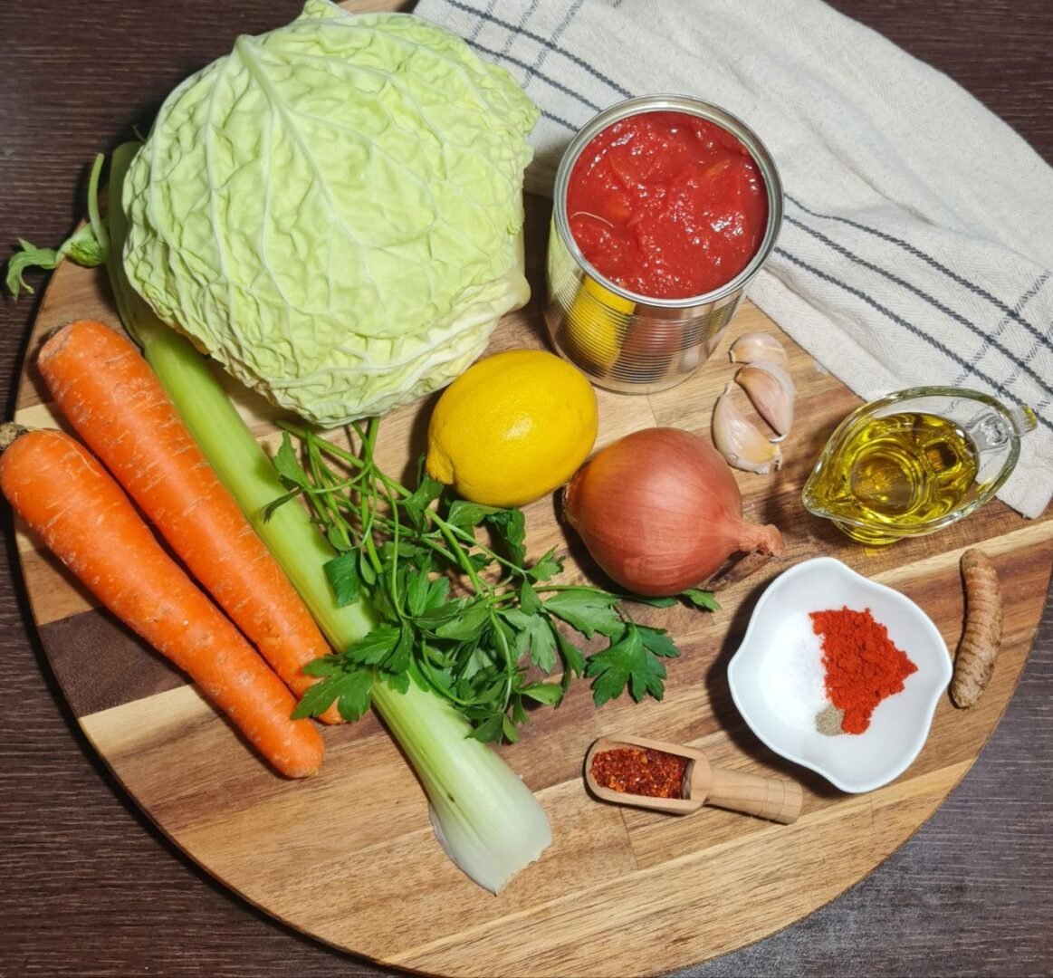  Ingredients for cabbage soup including cabbage, carrots, celery, lemon, onion, garlic, canned tomatoes, olive oil, parsley, spices, and turmeric on a wooden board.