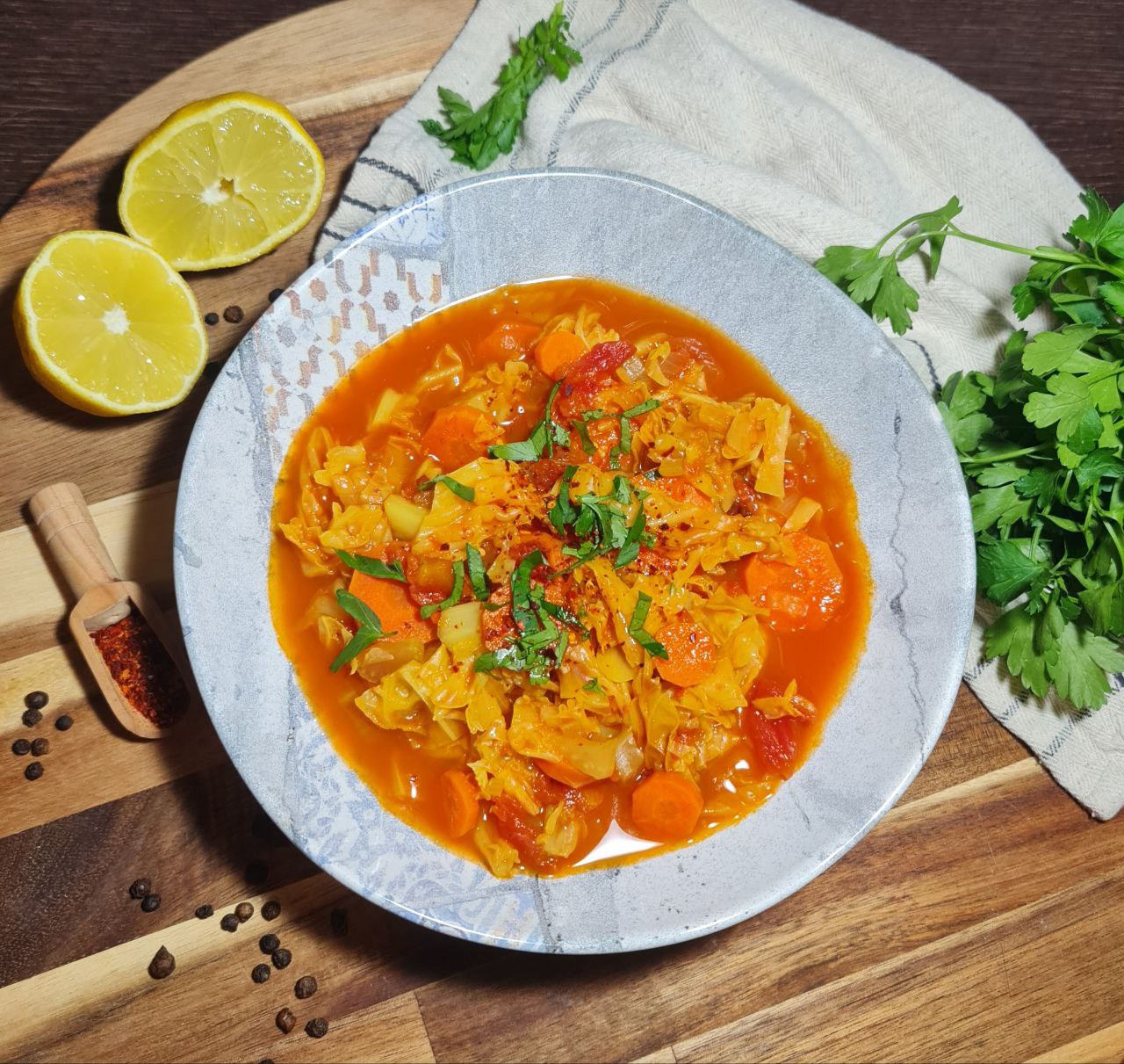 A bowl of vibrant cabbage soup garnished with fresh herbs, surrounded by lemon halves, parsley, and spices on a wooden board.