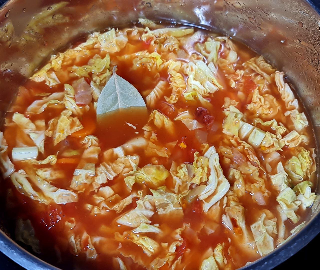 A pot of homemade cabbage soup with a bay leaf floating on top, featuring chopped cabbage, tomatoes, and a rich tomato-based broth.