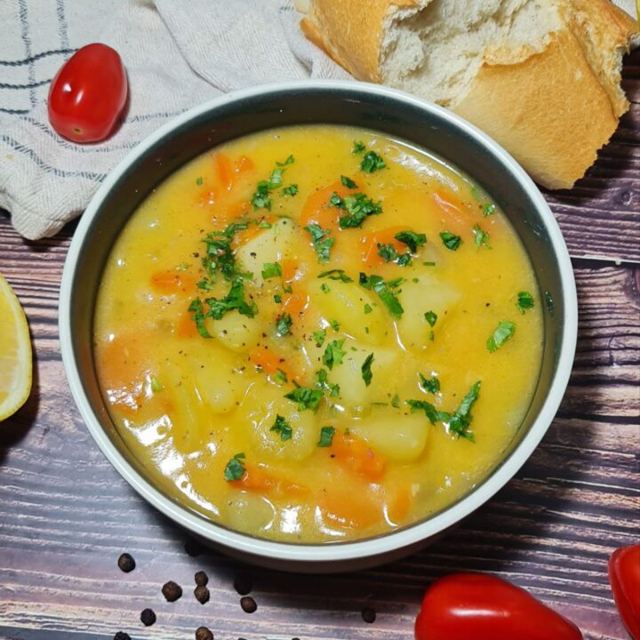 A bowl of carrot and potato soup garnished with fresh herbs, accompanied by a piece of bread, cherry tomatoes, and peppercorns on a wooden table.