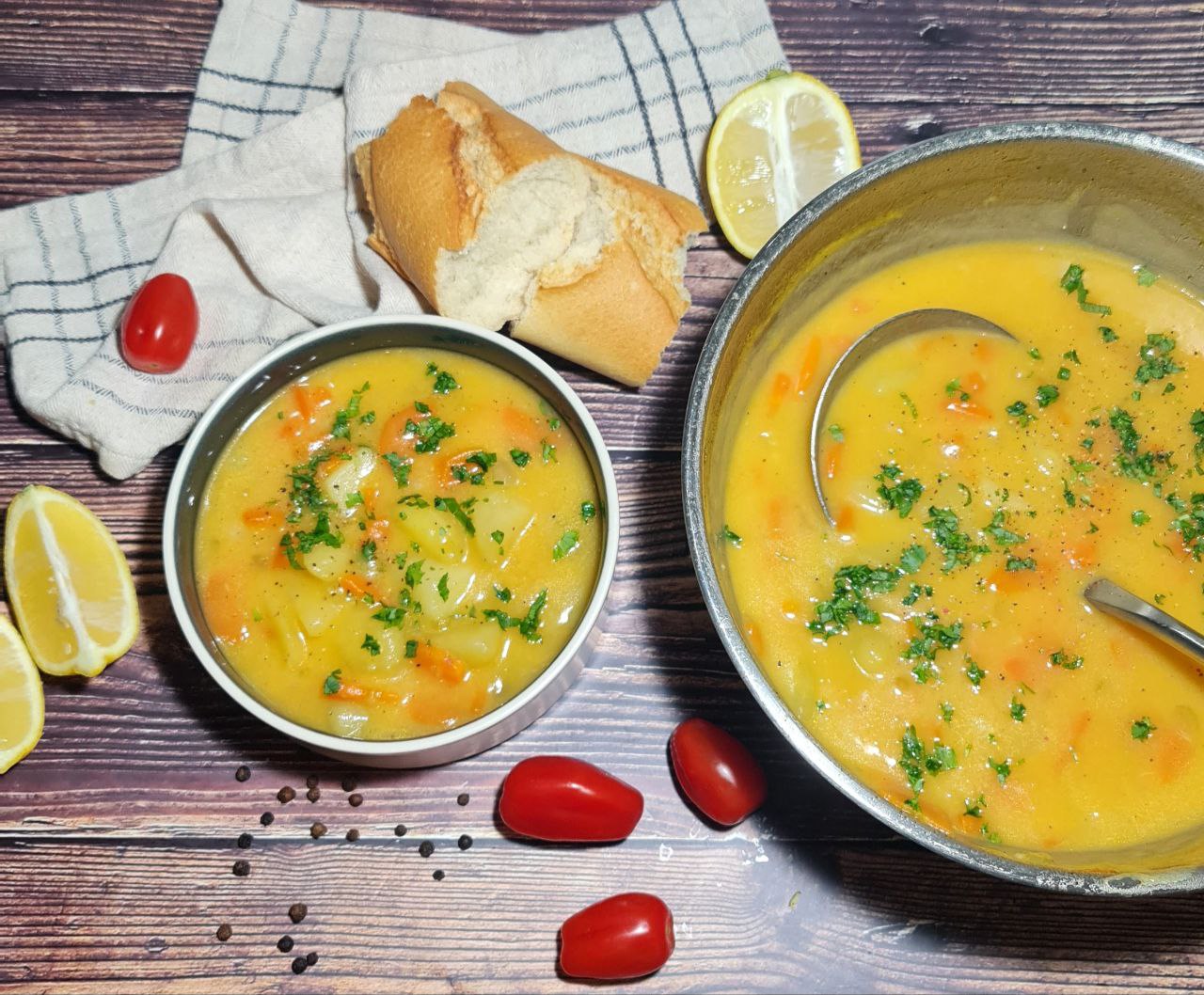 A bowl and pot of carrot potato soup garnished with fresh herbs, surrounded by lemon wedges, cherry tomatoes, black peppercorns, and a piece of bread on a wooden table.