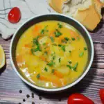 A bowl of carrot potato soup garnished with fresh herbs, accompanied by a piece of bread, cherry tomatoes, and peppercorns on a wooden table.