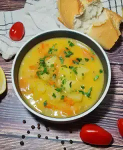 A bowl of carrot potato soup garnished with fresh herbs, accompanied by a piece of bread, cherry tomatoes, and peppercorns on a wooden table.