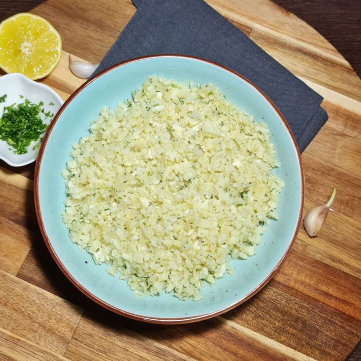 A bowl of freshly prepared cauliflower rice on a wooden board, accompanied by lemon, garlic, and chopped herbs.