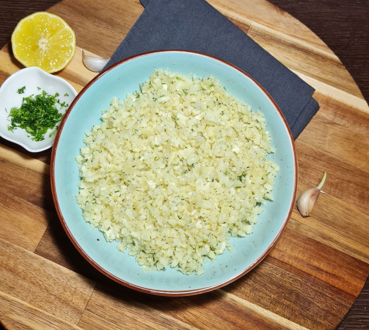 A bowl of freshly prepared cauliflower rice on a wooden board, accompanied by lemon, garlic, and chopped herbs.