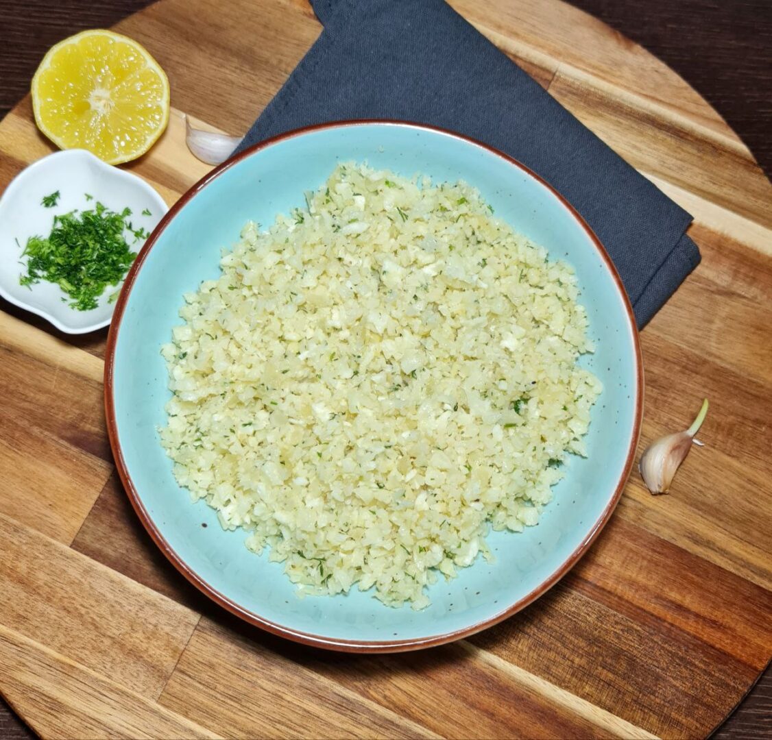 A bowl of freshly prepared cauliflower rice on a wooden board with lemon, garlic, and parsley garnish.