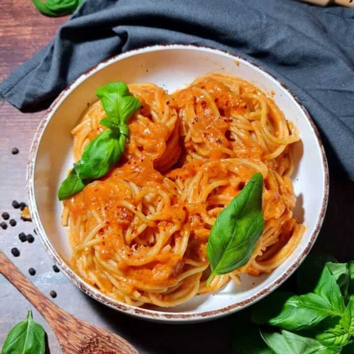 A bowl of spaghetti topped with a creamy coconut milk pasta sauce, garnished with fresh basil leaves, surrounded by ingredients like black pepper and red chili flakes.