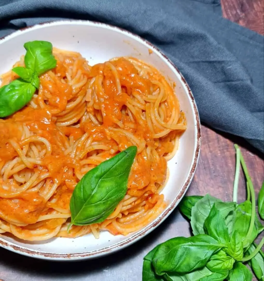 A bowl of spaghetti pasta topped with a creamy coconut milk tomato sauce, garnished with fresh basil leaves.