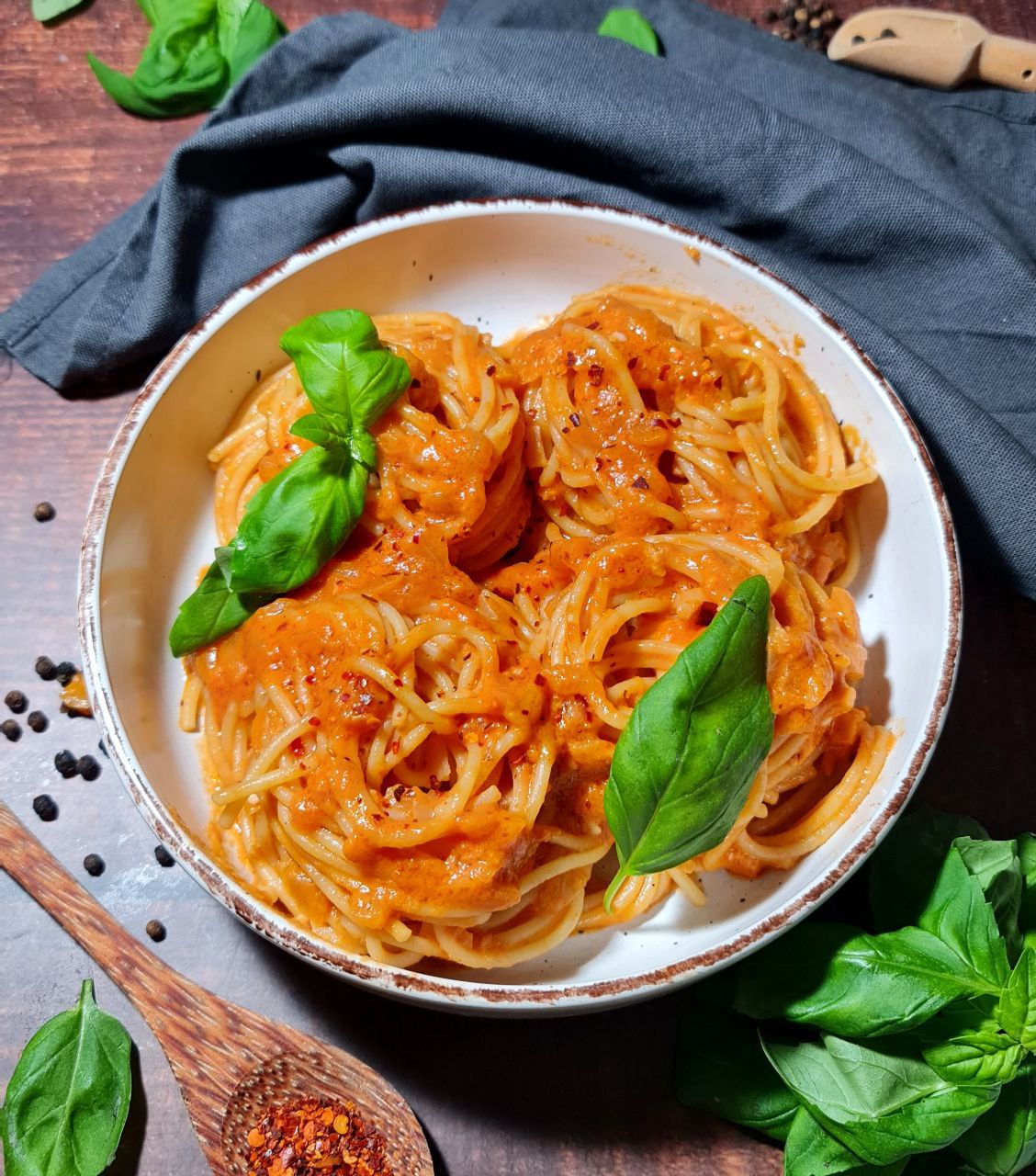 A bowl of spaghetti topped with a creamy coconut milk pasta sauce, garnished with fresh basil leaves, surrounded by ingredients like black pepper and red chili flakes.