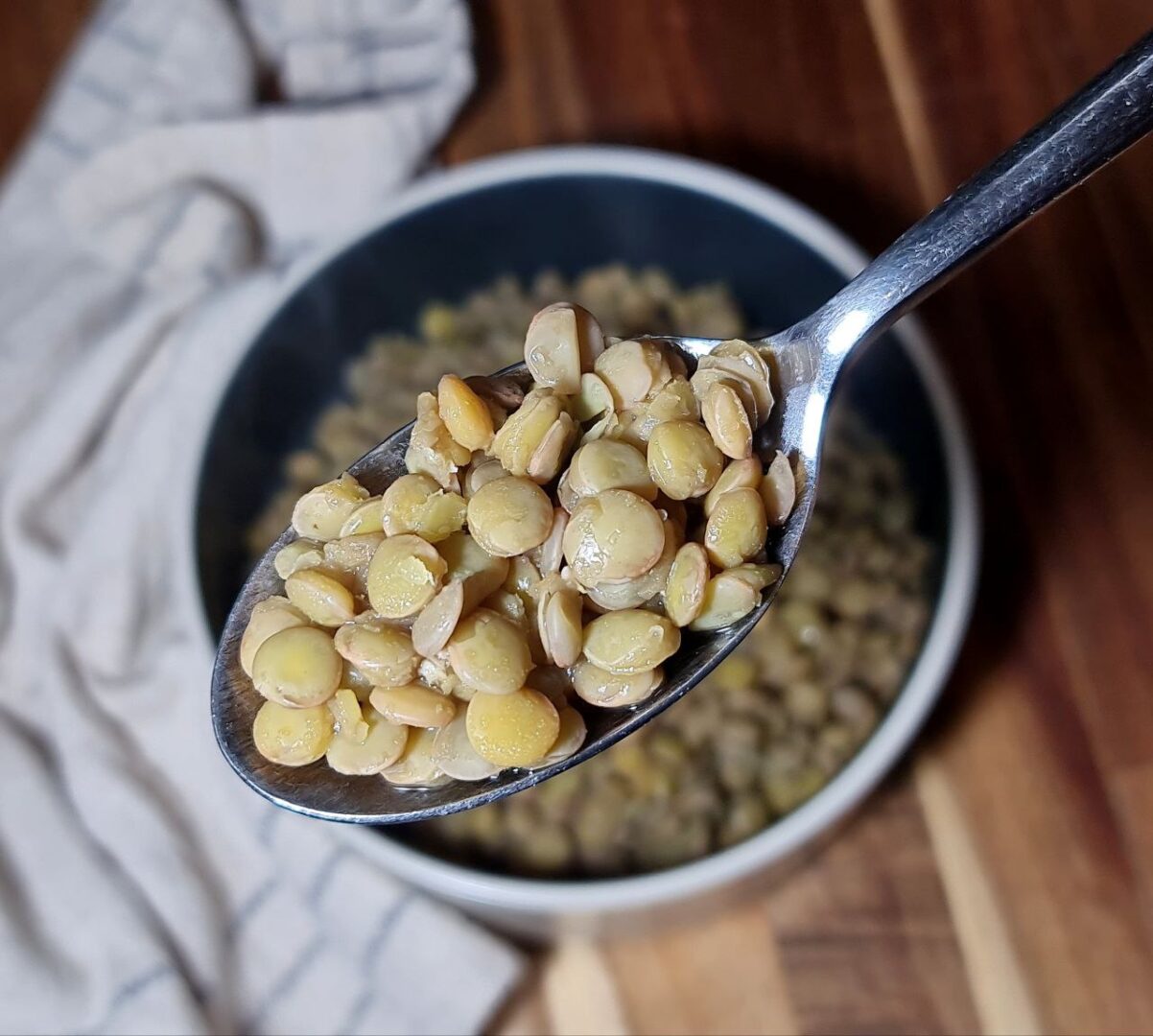 A close-up of a spoonful of cooked lentils held above a bowl of lentils with a cloth napkin in the background.