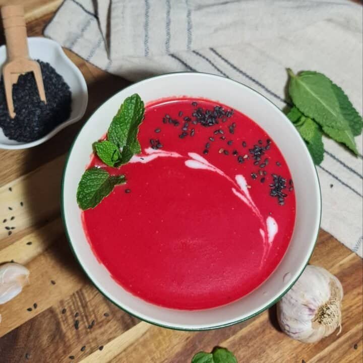 A bowl of creamy vegan beet soup garnished with fresh mint leaves and black sesame seeds, placed on a wooden surface with a small dish of black sesame seeds, garlic cloves, and mint leaves nearby.