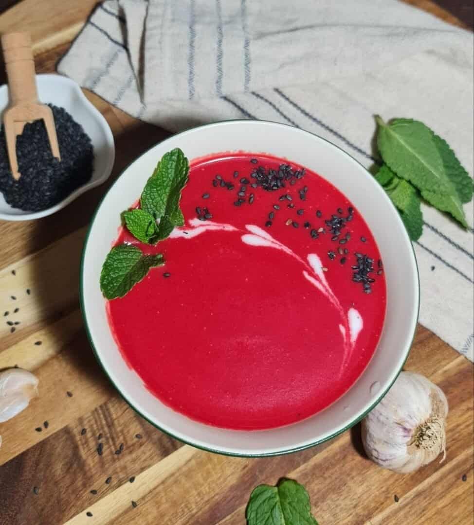 A bowl of creamy vegan beet soup garnished with fresh mint leaves and black sesame seeds, placed on a wooden surface with a small dish of black sesame seeds, garlic cloves, and mint leaves nearby.