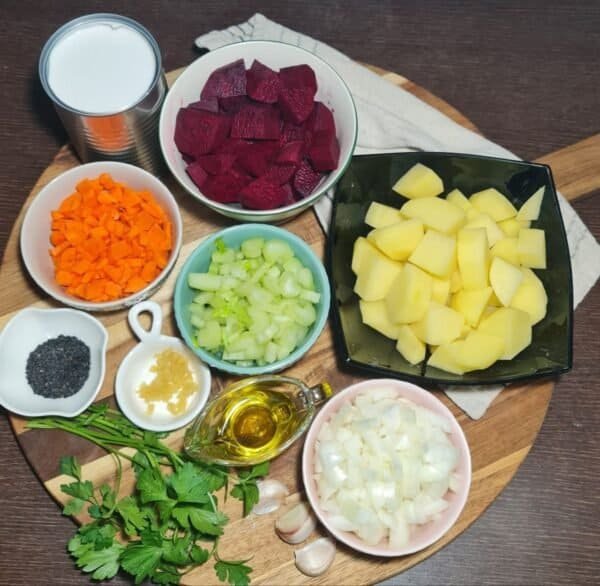 Ingredients for creamy vegan beet soup including chopped beets, potatoes, carrots, celery, onions, garlic, parsley, black sesame seeds, minced garlic, olive oil, and a can of coconut milk arranged on a wooden board.