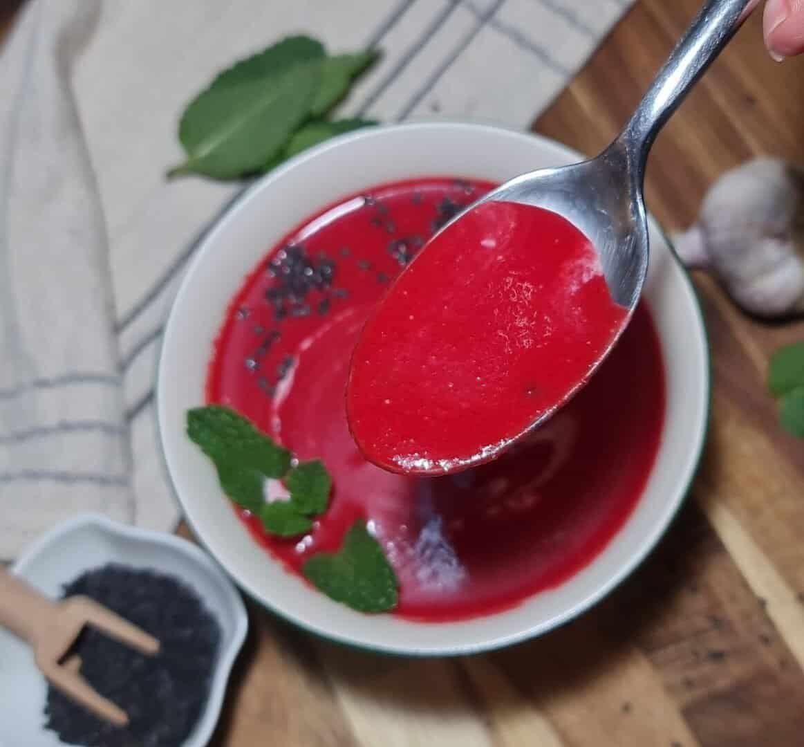 A bowl of creamy vegan beet soup garnished with mint leaves and black sesame seeds, with a spoonful of the vibrant red soup held above the bowl.