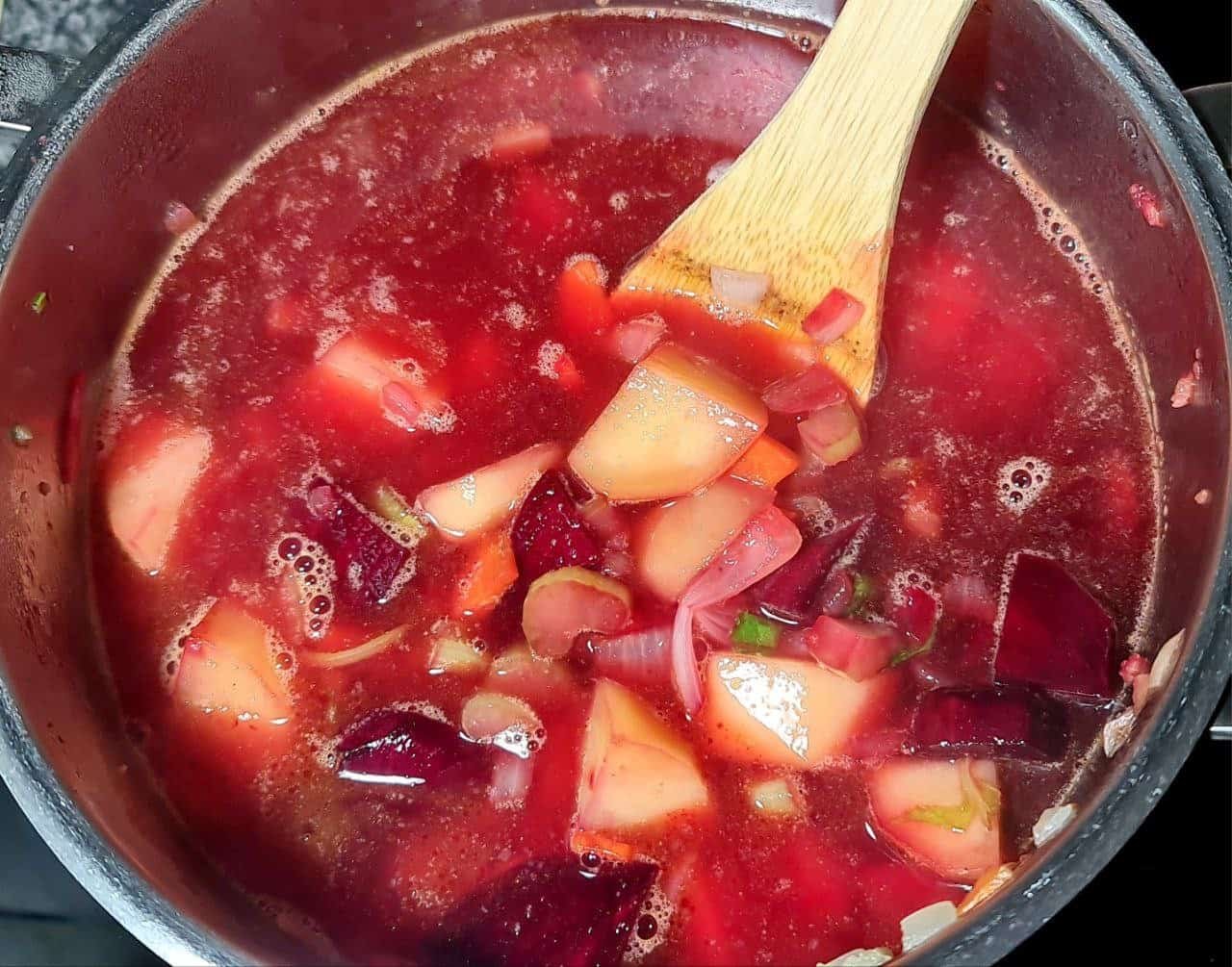 A pot of creamy vegan beet soup with chunks of beets, potatoes, and other vegetables being stirred with a wooden spoon.