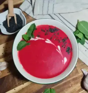 A bowl of creamy vegan beet soup garnished with fresh mint leaves and black sesame seeds, placed on a wooden surface with garlic cloves, mint leaves, and a small dish of black sesame seeds nearby.